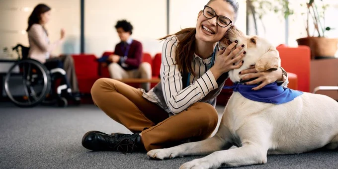 A light skinned woman, with glasses and her brown hair pulled back in a ponytail, snuggles with a large beige dog. The woman is smiling and facing the camera. She is wearing a white stripped long-sleeved shirt and brown pants. The dog is licking her cheek. In the background a woman in a wheelchair talks with a man who is sitting on red armchair. 