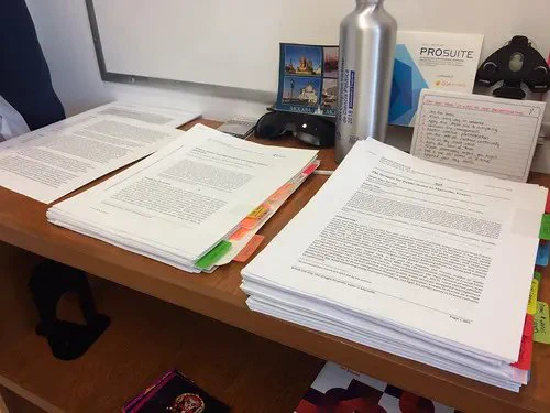 A set of three piles of printed articles on a wooden desk and a stainless steel water bottle.