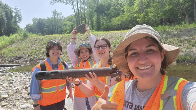 Four people with the WATER Institute pose for a selfie on a rocky stream bank. On the left, Denise Summers smiles. Next to her stands Sofie Liang holding a rock above her head and smiling. Third from the left stands Liz Hasenmueller smiling and holding the multiparameter sonde, a water quality instrument that looks like a black cylinder about 3 feet long and four inches in diameter. On the right stands Carly Finegan, smiling and giving a thumbs up with one hand and holding the camera with the other. Behind them is a shallow stream with a rising bank lined with trees and blue sky.