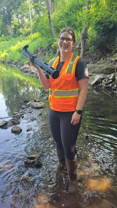 Liz Hasenmueller stands in the middle of a stream surrounded by green foliage, wearing tall rubber boots and an orange safety vest, holding the multiparameter sonde, a water quality instrument that looks like a black cylinder about 3 feet long and four inches in diameter. 