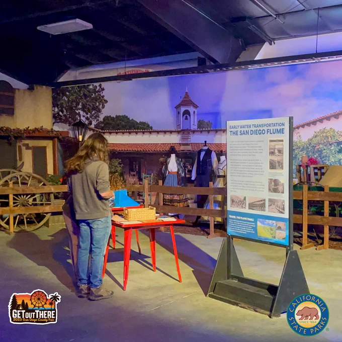 Image of a fair-goer looking at the California State Parks exhibit at the San Diego County Fair. There is a large backdrop photo of La Case Estudillo from Old Town State Historical Park. In front of that are mannequins dressed with period attire. There is a replica of an Adobe building to the left. There is a large text panel in the foreground whose title reads: Early Water Transportation. The San Diego Flume.