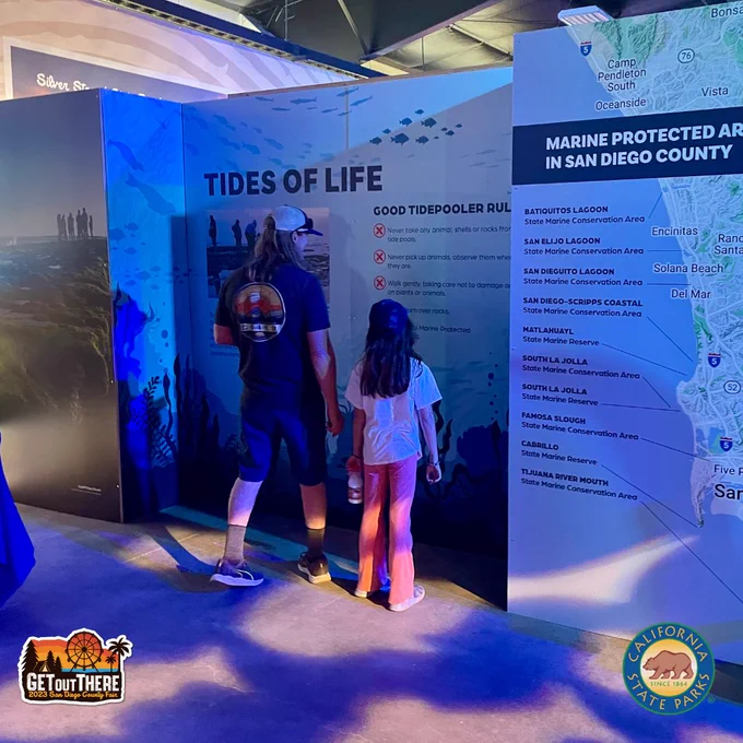 Image of two fair-goers reading the text on the wall of the California State Parks exhibit at the San Diego County Fair. The title of the largest panel reads: Tides of Life. The title of the secondary panel reads: Marine Protected Areas in San Diego County.