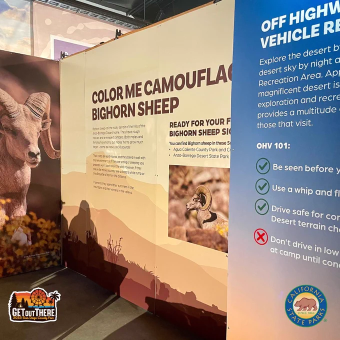 Image of large text panels of the California State Parks Exhibit at the San Diego County Fair. There is a large image of a big horn sheep to the left. The large text panel in the middle is yellow and tan and has a silhouette  of a desert landscape. The large title on that panel reads: Color me Camouflaged-Bighorn Sheep. The smaller text panel to the right of that is blue and the title reads: Off Highway Vehicle Recreation Area.