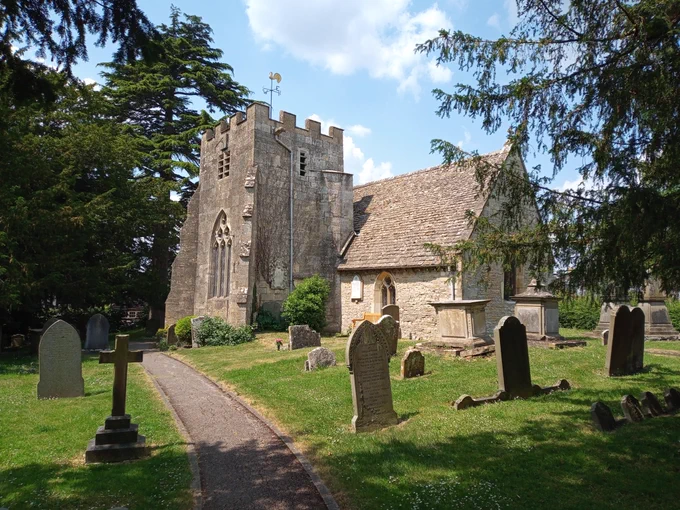 The Church of St Catherine, Staverton, Gloucestershire. Photo taken from the southeast of the church, showing the Chancel to the right and a short, square plan tower to the left, with gravestones in front of both. The church walls are made of limestone rubble stonework, and the roofs are also made of limestone slates; the top part of the tower is crenelated. The gravestones are set in a grassed churchyard with an access path running from the centre bottom of the photo past the left side of the tower. Overhanging trees frame the photo on the left and right sides.