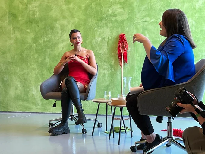 Two women sit on grey chairs in a green-washed gallery space. 