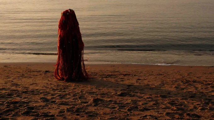 A film still shows a figure dressed in red fabric stands at the shore on a beach at sunset. 