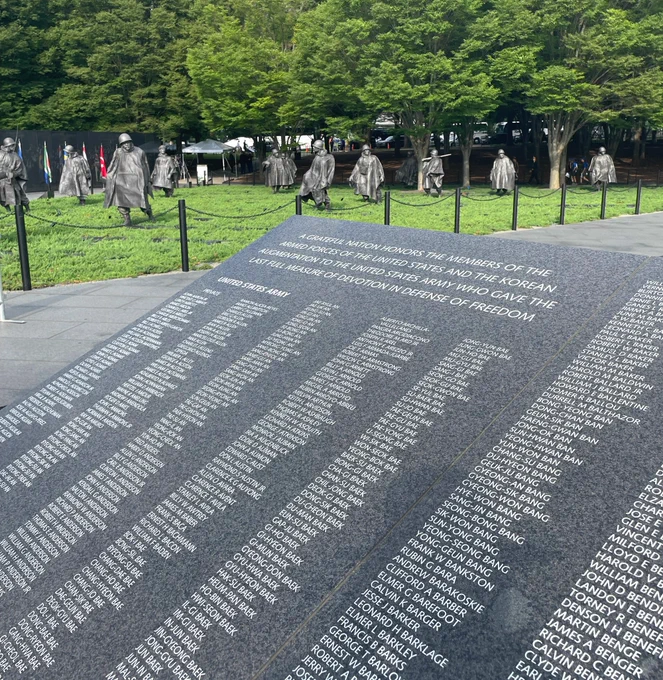 A large gray stone tablet covered in lines of names stands in an outdoor military memorial with a group of large metal statues of American soldiers standing on a lawn behind it.