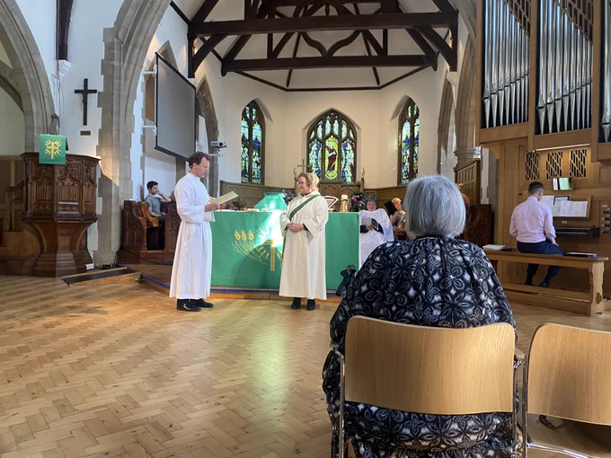 Rev Brutus welcoming Rev Sarah during the service and standing in front of the altar with the choir seated behind and an organist sitting at the organ to one side. Both are wearing white robes.