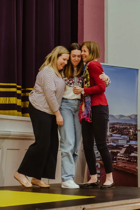 three women embrace on stage. They all smile.