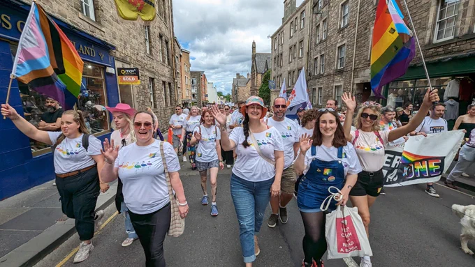 Group of Edinburgh Zoo and RZSS staff walking in the parch at Pride wearing white t shirts with rainbow flamingos and a rainbow Edinburgh Zoo logo on them. Staff on the left and right are holding pride flags.