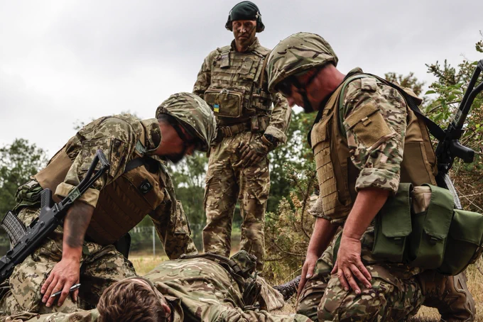 A British Soldier in combat uniform from the Rifles Regiment oversees two Ukrainian soldiers in combat uniform conducting battle casualty first aid on a third Ukrainian soldier pretending to be injured.