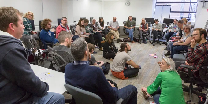 People are sitting in a circle inside a meeting room. Some are on chairs, others are on the floor.