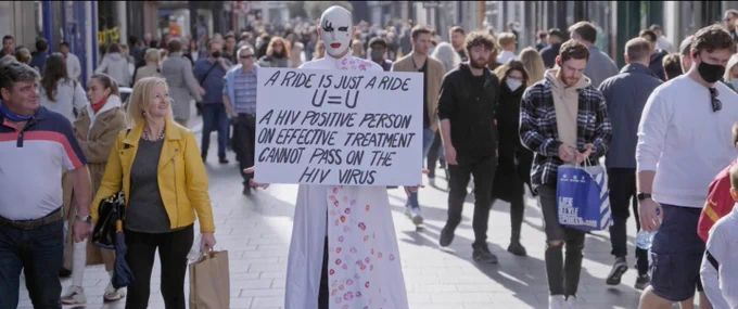 An person dressed all in white stands in the middle of a busy street with a sign that reads: A ride is just a ride. U=U A HIV positive person on effective treatment cannot pass on the HIV virus. They have no hair visible and their face is painted all white, with red lips and a graphic black design on one eye.
