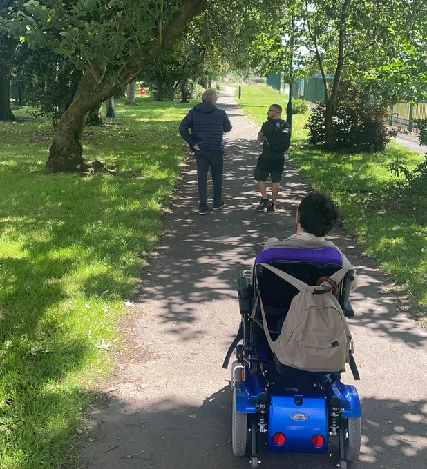 Seb, a dark haired person in a wheelchair, wheels through sunny, Bolton Road Park in Bury. Two people walk ahead on the path.