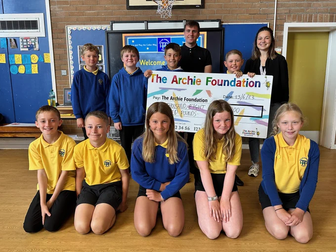 Cullen Primary School pupils in their uniform of yellow polo shirts and blue sweatshirts, gather around an oversized cheque which they are presenting to the Archie Foundation.