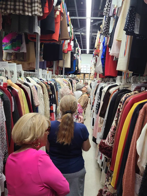 A group of senior adults walk down an aisle between two racks of fashion artifacts. They are guided by a collection staff member.
