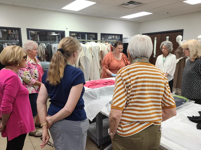 A group of senior adults stand around a table containing an article of clothing. A collection staff member discusses the artifact.