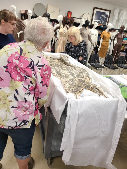 A group of senior adults stand around a table containing an article of clothing. A collection staff member discusses the artifact.