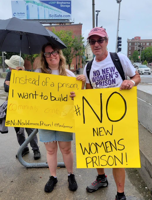 A blonde white woman and white man wearing a baseball cap hold yellow signs. The woman sign needs instead of a prison I want to build community centers hashtag no new women's prison! At Max governor the man sign reads no new women's prison in very large handwriting