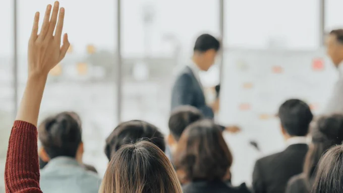 Image of a person raising their hand in the audience of an event where two people are presenting in a blurred background.