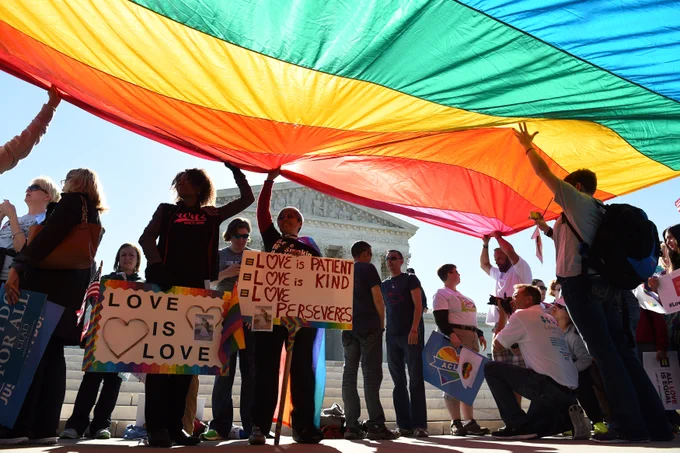 A rainbow flag being held up by people in front of the U.S. Supreme Court.