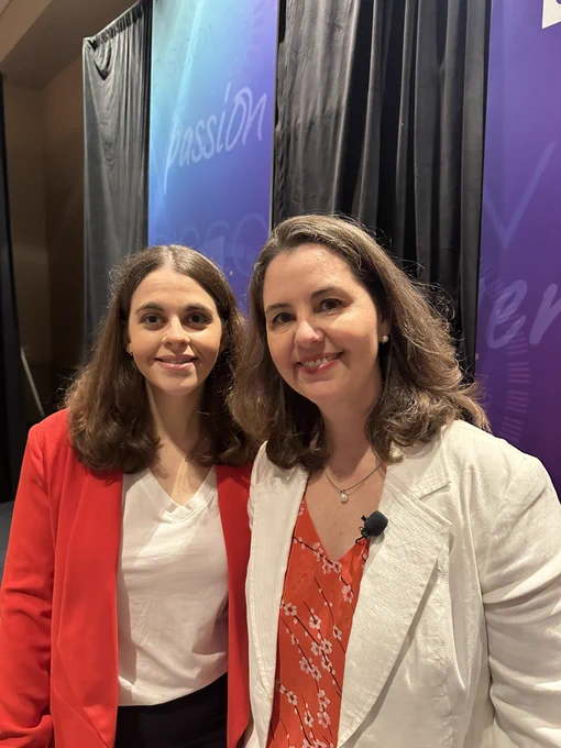 Jordyn wearing a red blazer and white shirt, Janette wearing a white blazer and orange shirt, standing together on the ISTE stage.