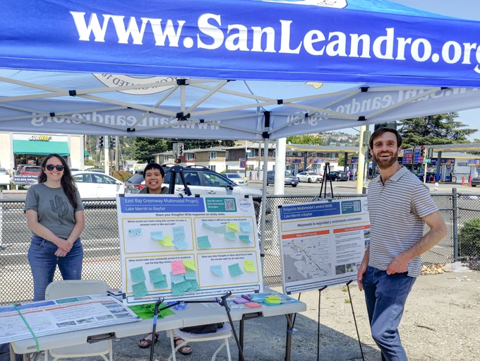 Two people stand in front of a booth with a banner that reads "www.SanLeandro.com"