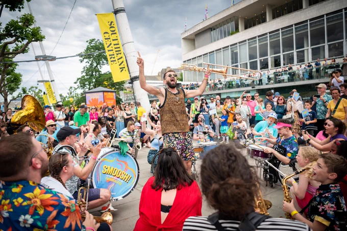 Outside, the Southbank Centre to the right, banners above read Southbank Centre in the organisations brand colour and font. A hug crowd are gathers in a circle around musicians crouched playing percussion, brass, and woodwind. In the centre a man in sunglasses and loud clothes holds a trombone, his arms wide he points to the sky, his mouth open with joy as he encourages the crowd. 

