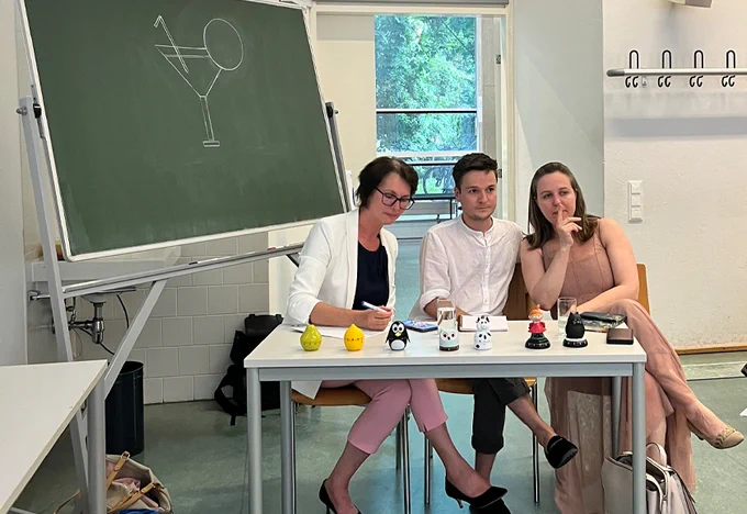 Die Jury, zwei Frauen und ein Mann sitzen an einem weißen Tisch; vor ihnen stehen sechs unterschiedliche Eieruhren. Links hinter ihnen steht eine grüne Tafel, auf die ein Cocktailglas gezeichnet ist.
