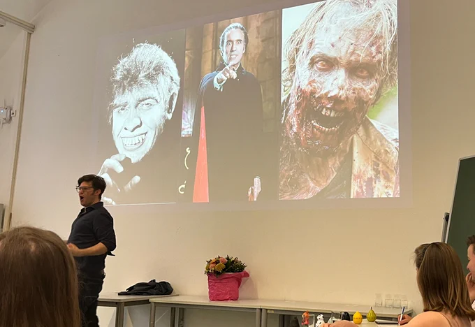 Teilnehmer des Humanities Slam trägt im Stehen vor Publikum vor. Hinter ihm eine Power Point slide mit drei Horrorportraits.