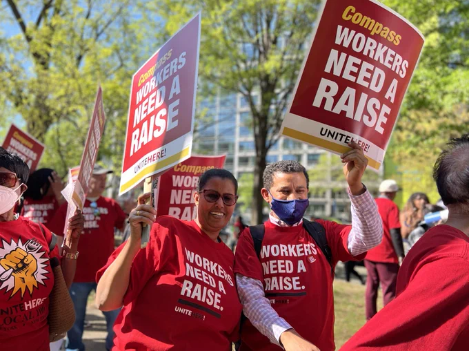 Workers in red shirts that read "Workers Need a Raise." They are walking in a picket line, smiling and holding "Compass Workers Need a Raise" signs.