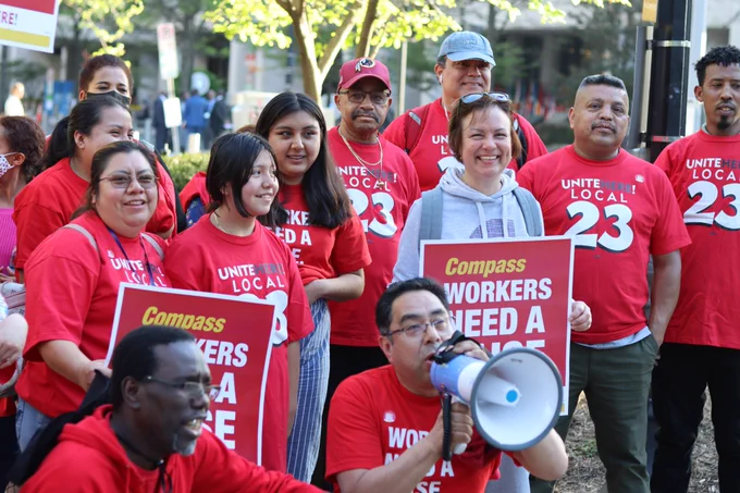 A group of workers and their families pose for a photo. They are wearing red UNITE HERE Local 23 tshirts and holding signs that say "Compass Workers Need a Raise." 