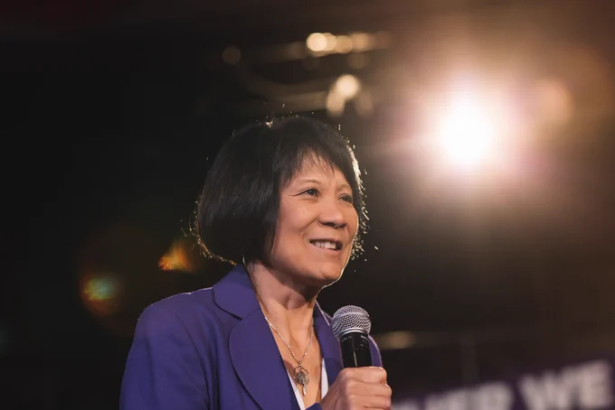 Olivia Chow has a chin-length hair, and is wearing a purple coat. She's holding a microphone.