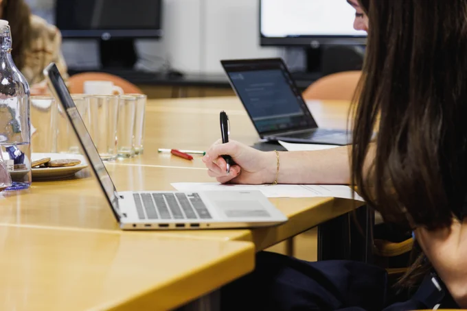 photograph shows a young woman working at a desk in front of her laptop. She is holding a pen in her right hand, ready to start writing.