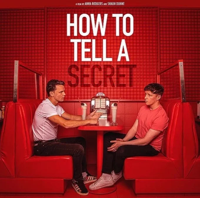 HIV Activist Robbie Lawlor and director Shaun Dunne sit in a colour blocked red restaurant booth. The title of the movie How To Tell A Secret is written on the wall between them. They both look serious as Robbie has a secret he needs to tell the film director and former lover Shaun.