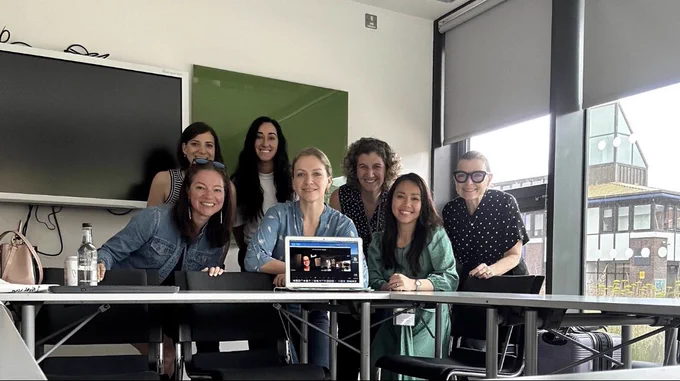 Group of women in a classroom standing together smiling with a laptop.