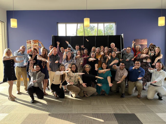 A group of 34 UF CALS faculty and staff posing with gator chomp hand motion for a group photograph in a large room. 