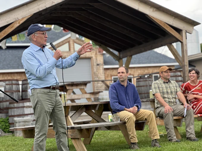 Picture of Peter standing and speaking into a microphone outside while others look on. 