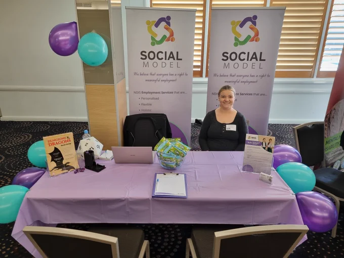 A photo of a siling woman in a black top sitting at an expo stall.
There are purple and teal balloons around a table with a purple tablecloth.  There are flyers, business cards, and popcorn on the table.
Behind the woman are two banners that read "Social Model.  We believe that everyone has a right to meaningful employment."