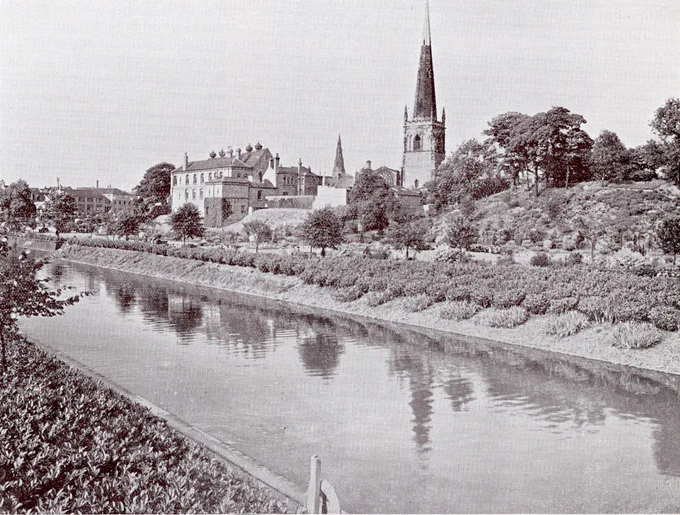 Black and white photograph of a public garden with view of the river in the foreground and a church and great hall in the background.