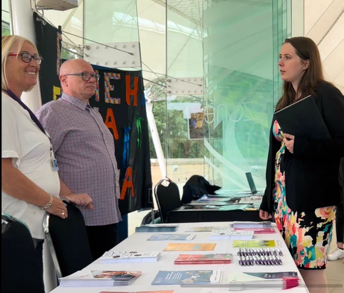 Emma stands speaking to two people at the event. There is a table between her and the two other people. One the table are many piles of information booklets and some pens. 