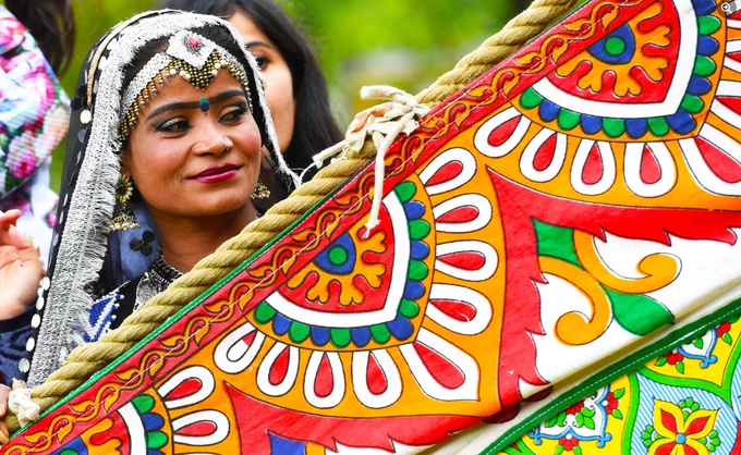 person with ornate headdress holding a vibrantly patterned item looking to the side in an outdoor park setting.