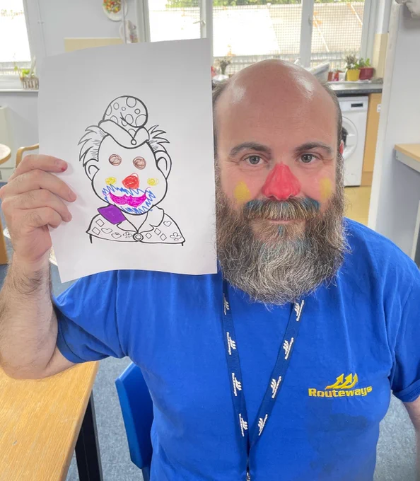 Aaron, a bearded man in his 40s smiles at the camera. He's holding a picture with a clown make up design on. On his face is face paint of a similar design.