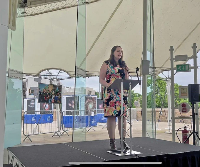 Emma stands on a small stage at the Dynamic Earth Science Centre in Edinburgh. She is standing in front of a lectern speaking to the audience who are behind the camera. There are large floor-to-ceiling glass panels behind her allowing a view of outside where there are large science centre information pop-up cubes, some metal railings and some green trees and bushes. 