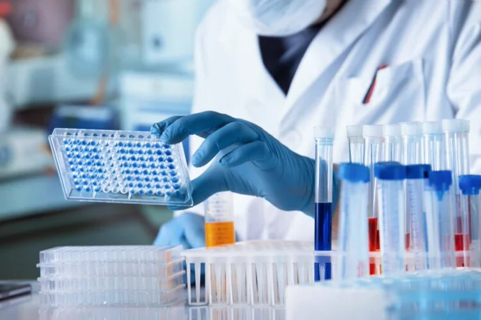 Researcher in a lab wearing blue latex gloves holding a small rectangular plate with wells in containing samples. A tray of test tubes are in the bottom right corner.