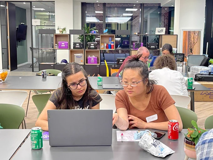 An office setting. Two women in the foreground are looking at a shared laptop. Three people are visible in the background.