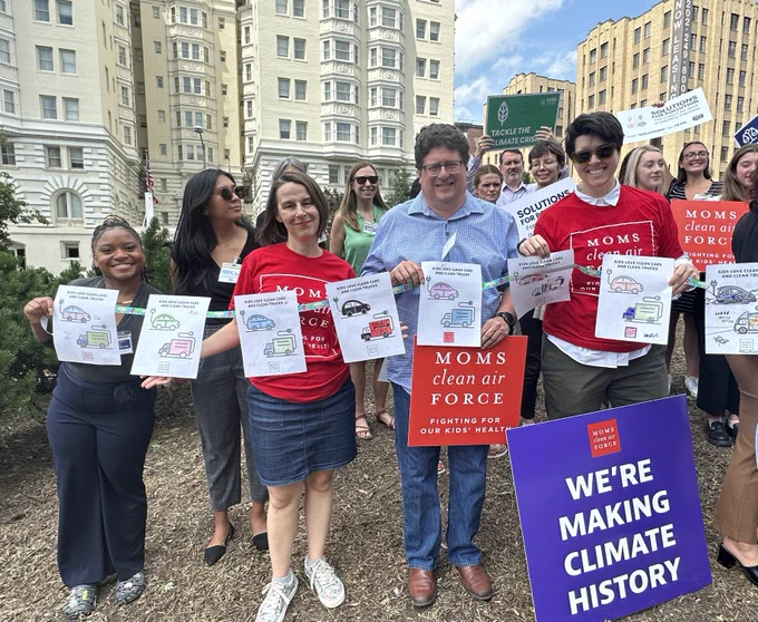 Moms in red shirts and their partners hold up coloring book pages of electric vehicles.