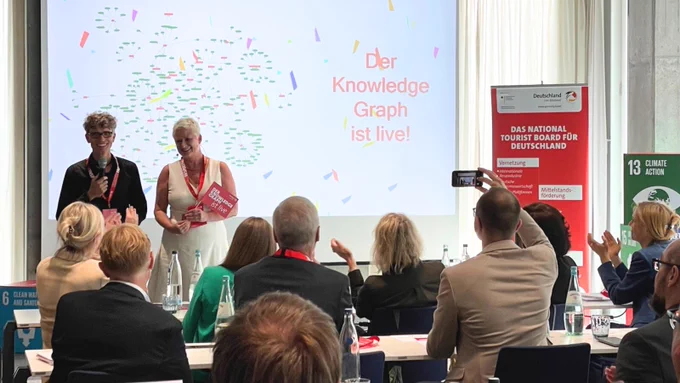 Launch des DZT Knowledge Graphen: Im Hintergrund sieht man auf einer Leinwand eine Präsentation mit dem Text "Der Knowledge Graph ist live!" Im Vordergrund sitzt das Publikum. 