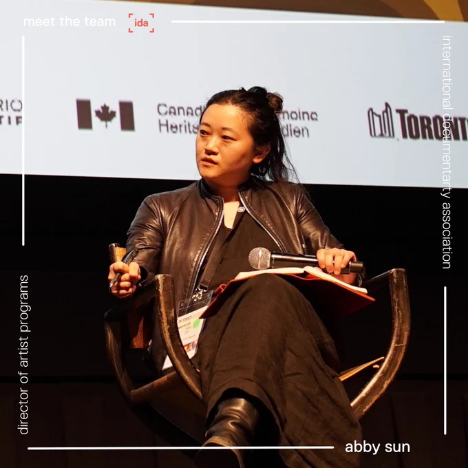 Flyer for IDA Meet the Team campaign. Meet Abby Sun. Director of Artist Programs. Image of a woman of Asian descent, sitting in a brown chair on a stage with her hair in a messy bun, wearing a black jacket, shirt, and pants. 