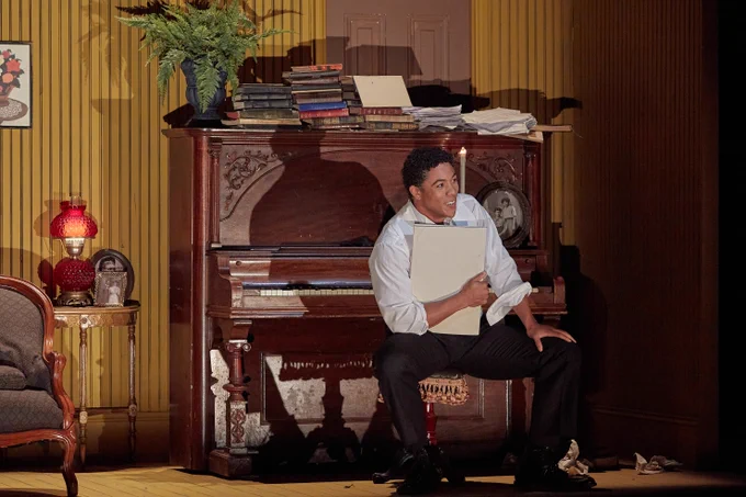 A man sits on a piano bench, facing away from the piano, holding a score under his arm